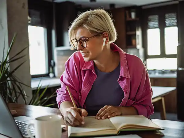 Young woman with glasses looking at laptop and taking notes. She is working at home.